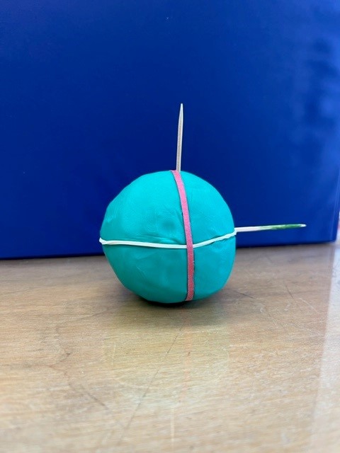A blue clay ball with two rubber bands around its axis and a stick through it.